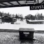 Ett svartvitt foto från en tågperrong. Skylten i perrongens tak lyder: STUVSTA. Ett flebostadshus, några trälador och en villa syns bland snön.