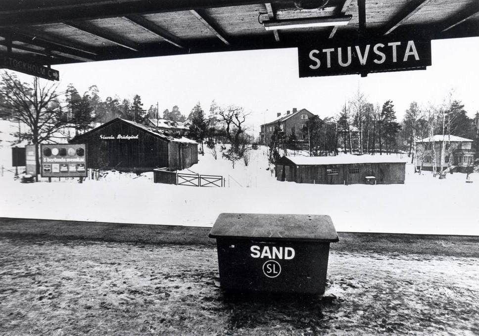 Ett svartvitt foto från en tågperrong. Skylten i perrongens tak lyder: STUVSTA. Ett flebostadshus, några trälador och en villa syns bland snön.