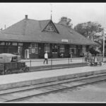 Svartvitt foto av stationshuset med två tågspår och perrong i förgrunden. En dressin som ser ut som en T-ford står på spåret.