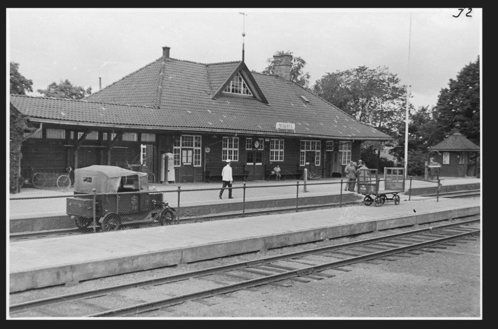 Svartvitt foto av stationshuset med två tågspår och perrong i förgrunden. En dressin som ser ut som en T-ford står på spåret.