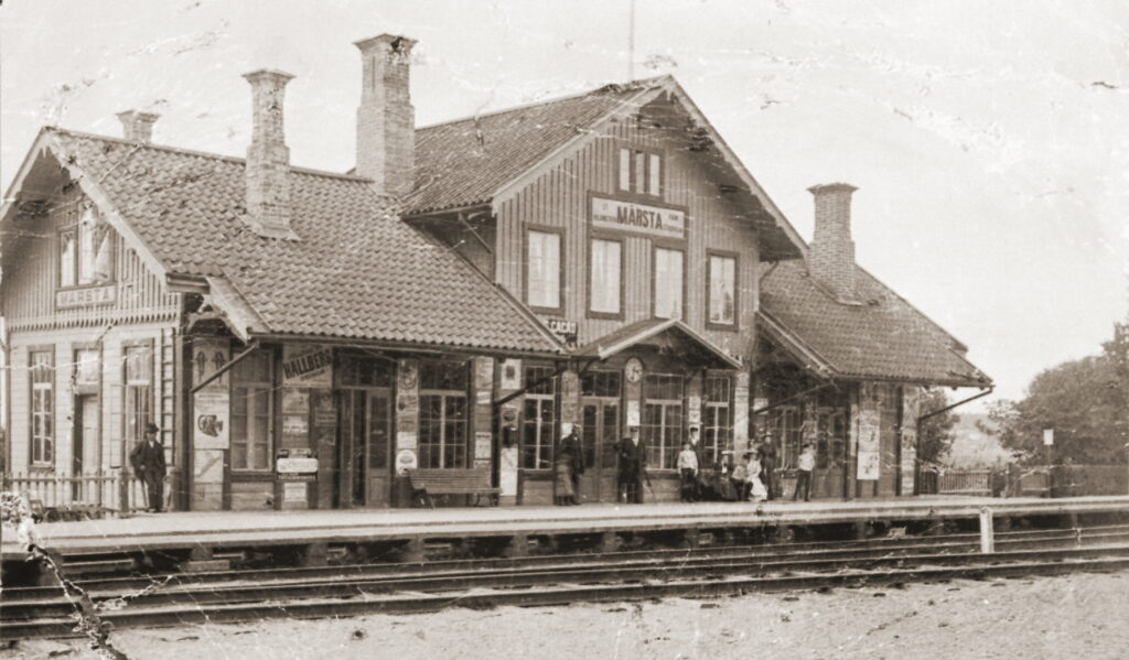 En svartvit bild av stationshuset med väntande resenärer.