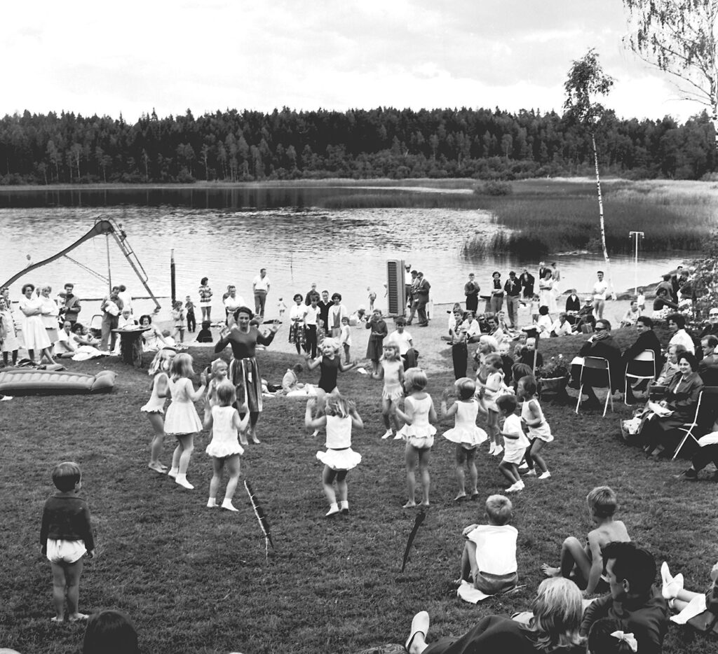 Barn dansar i cirkel på en badstrand med publik.