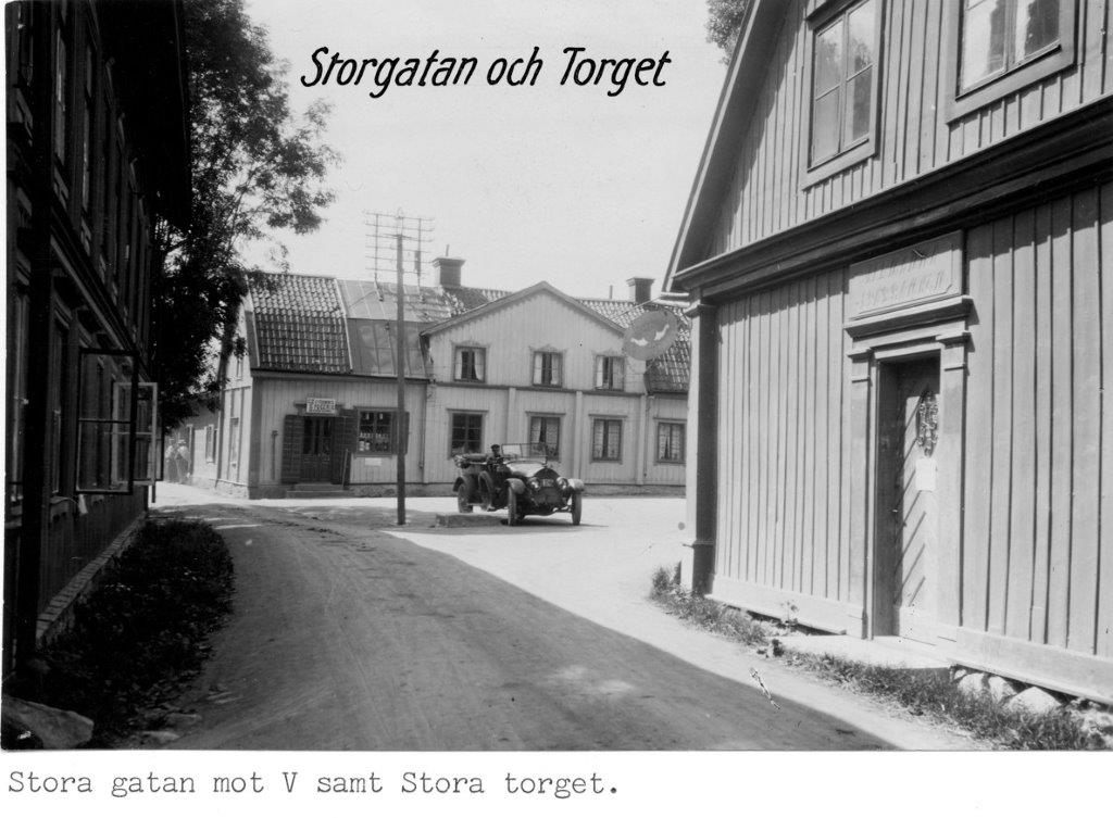 Ett svartvitt vykort. Mellan husen öppnar sig torget där det står en gammaldags öppen bil med en förare.