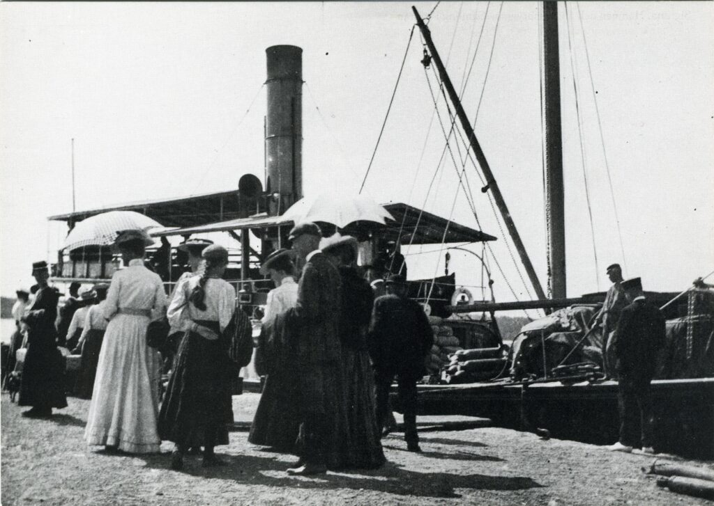 Män och kvinnor, varav två med parasoller står i solskenet på bryggan där en ångbåt ligger förtöjd och lastad med varor.