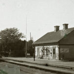 Ett svartvitt foto av ett stationshus där ett ånglok kommer in på perrongen. En ensam man står på perrongen och i bakgrunden syns ett enda annat hus.