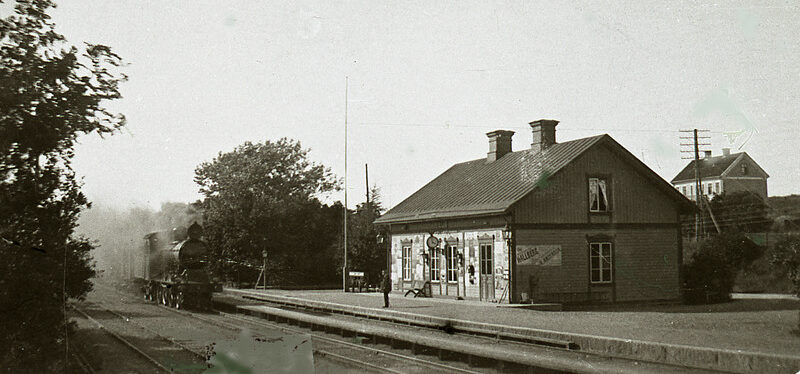 Ett svartvitt foto av ett stationshus där ett ånglok kommer in på perrongen. En ensam man står på perrongen och i bakgrunden syns ett enda annat hus.