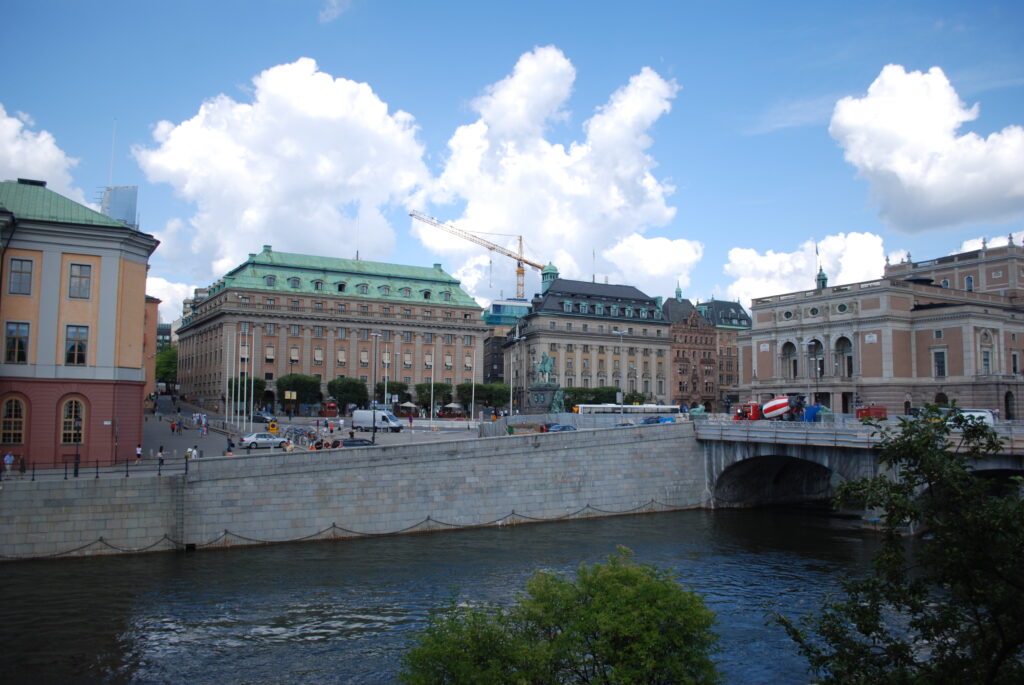 Ett färgfotografi av torget i modern tid med operan till höger och andra äldre hus runt toget. I förgrunden ses Norrströms vatten.