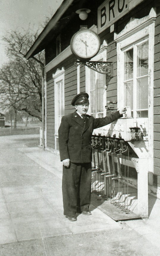 Ett svartvitt foto av en stins i uniform som står under en klocka som vilar på en sirlig gjutjärnsarm ut från stationsbyggnaden.