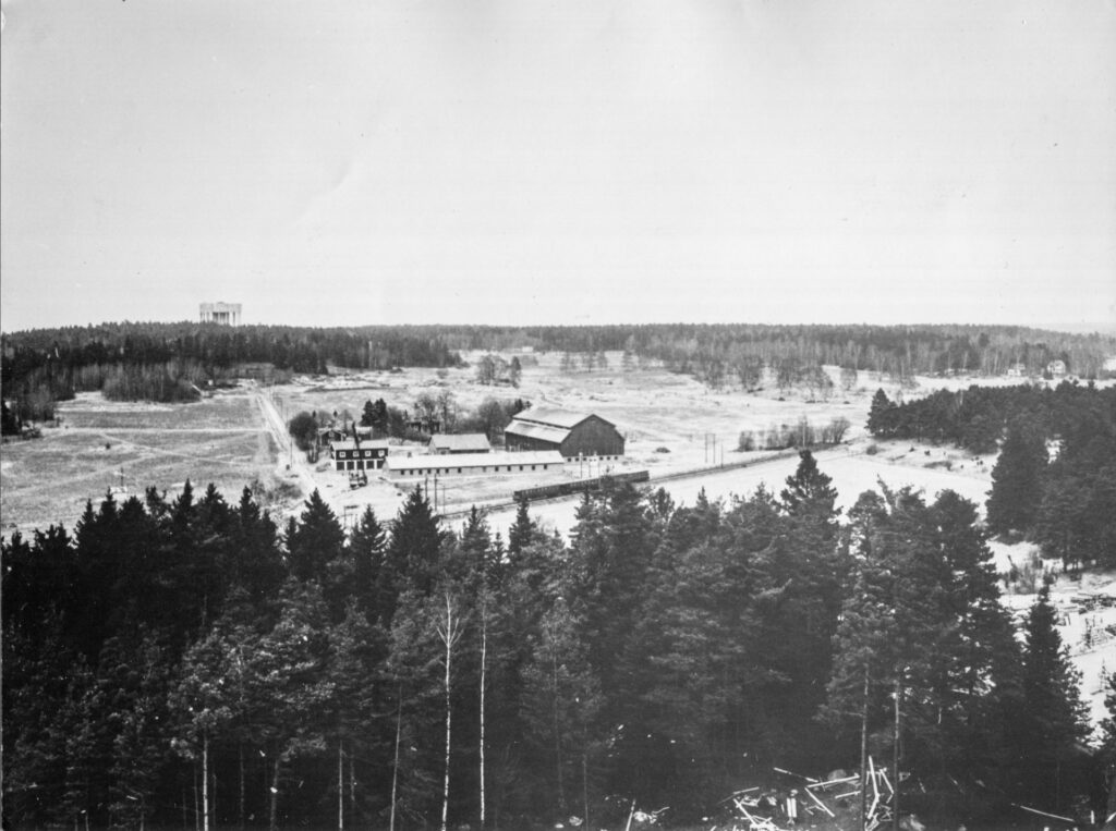 Ett svartvitt foto visar en gård med stor lada mitt på en äng som öppnar sig mellan skogen i för- och bakgrunden.