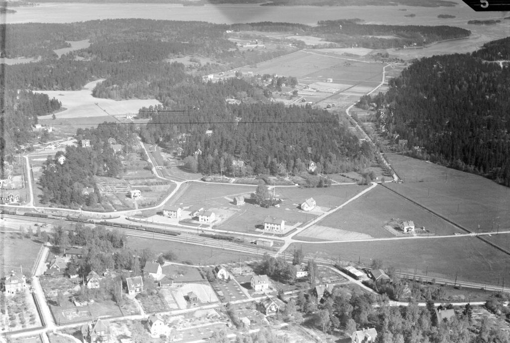 Svartvitt flygfoto med järnväg som går genom ett öppet landskap med enstaka villor.