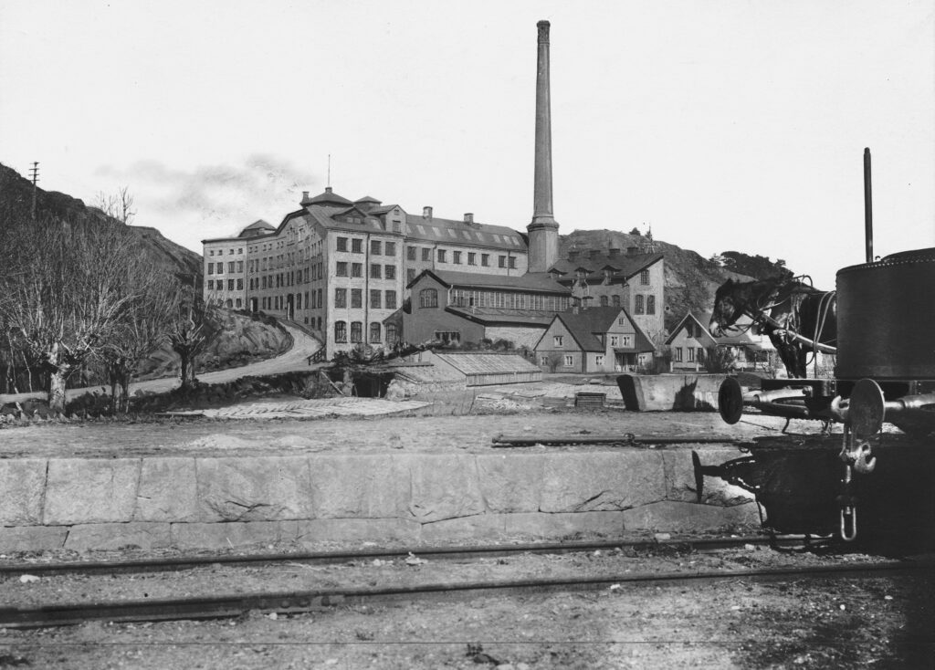 Ett svartvitt foto av en fabriksbyggnad på håll. Järnvägsspår och del av en vagn i förgrunden.