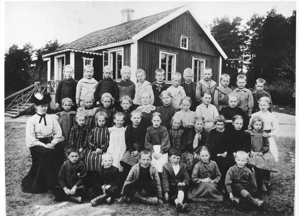 Ett svartvitt foto av en lärarinna i hatt med sina skolbarn framför den lilla skolbyggnaden.