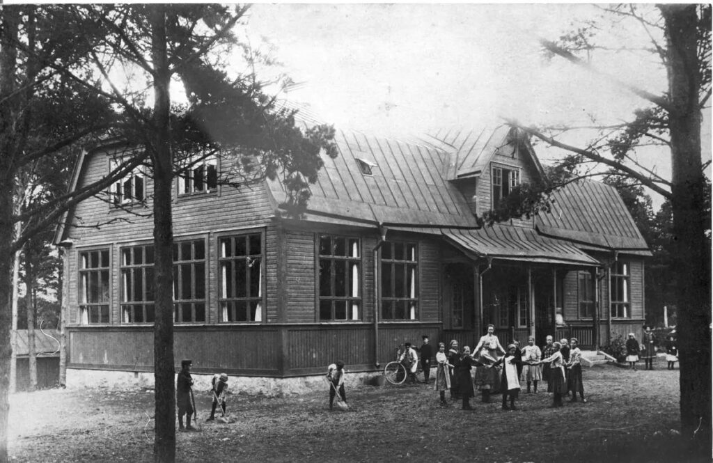 Ett svartvitt foto av en träskola där några pojkar gräver på skolgården och en kvinna står i en ring tillsammans med en grupp flickor som tittar mot kameran.