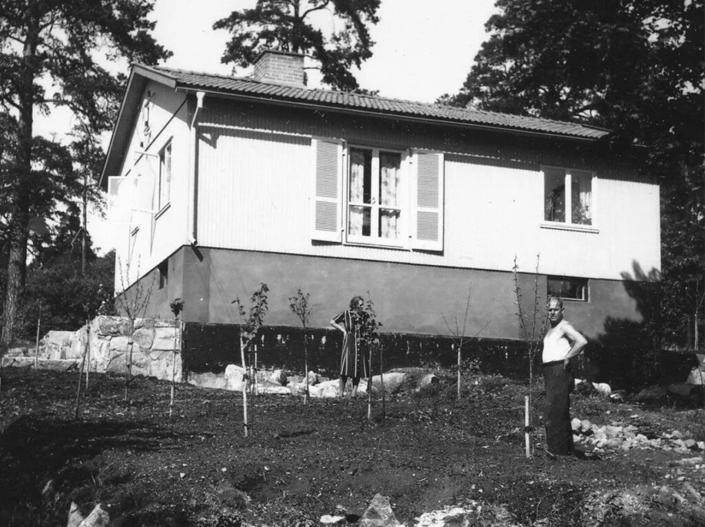 Ett svartvitt foto av ett litet hus. En man och en kvinna står bland nyplanterade träd.