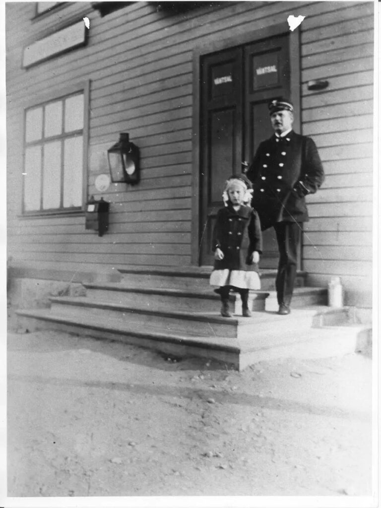 Svartvitt foto. Den lilla flickan står tillsammans med sin uniformsklädda pappa på trappan till stationshuset. På dörren bakom dem står det "väntsal".