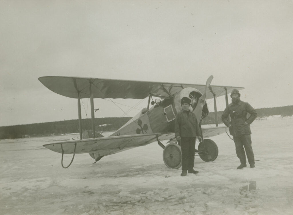 Två män står på isen framför ett flygplan med dubbla vingar och propeller.