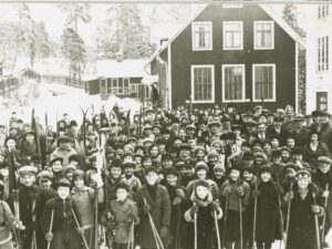 Ett hundratal barn står uppställda med vinterkläder och skidor framför skolhuset.