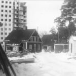 Svartvit bild där ett högt punkthus under byggande sticker upp bakom två äldre trähus. vinter.